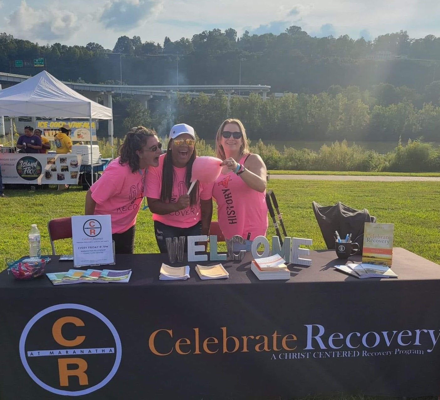 three women laughing behind table reading "Celebrate Recovery: A Christ Centered Recovery Program"